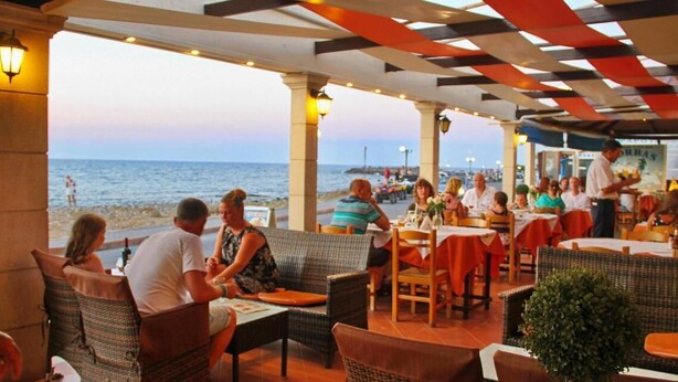 An outdoor dining area with wicker furniture, tables with red tablecloths, and a view of the sea. - Kalia Beach (Hersonissos)