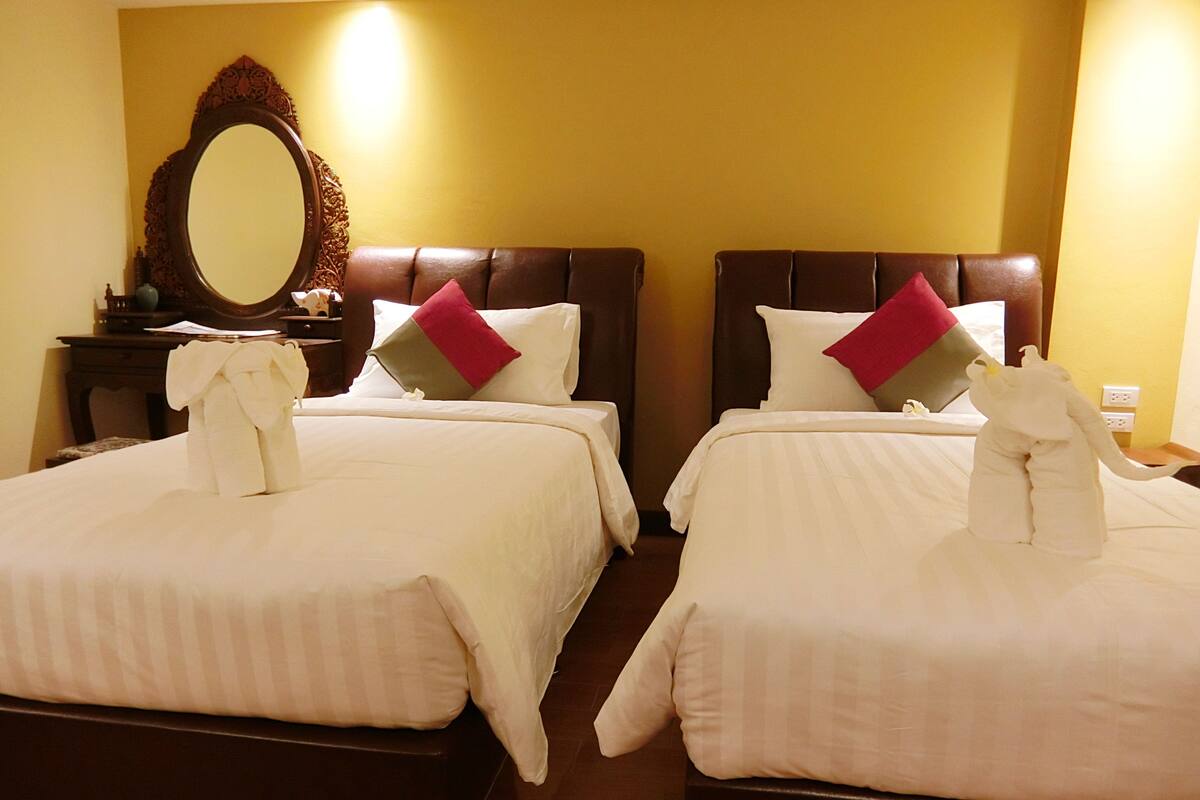 Deux lits doubles avec du linge de lit blanc et des oreillers rouges et gris, une tête de lit en bois, un miroir et une petite table avec une lampe.