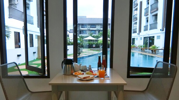 Mesa de desayuno con cafetera, tetera y jugo, con vista a una piscina y un patio con plantas y sombrillas.