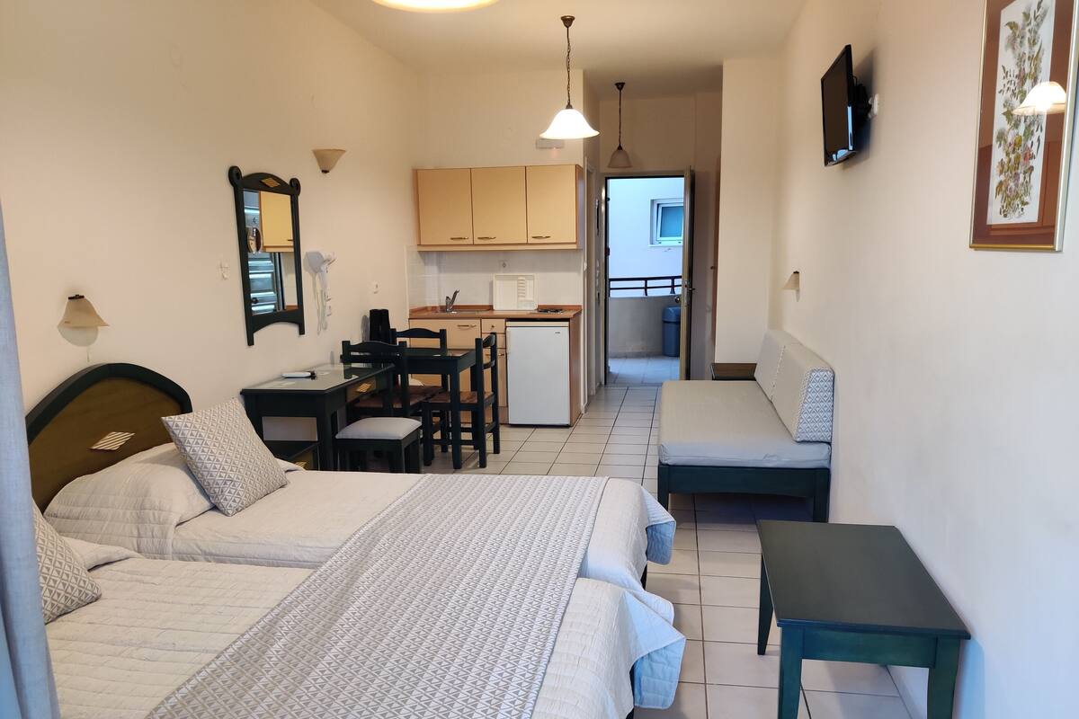 A compact hotel room with two beds, a small kitchenette, a dining area, and a balcony.