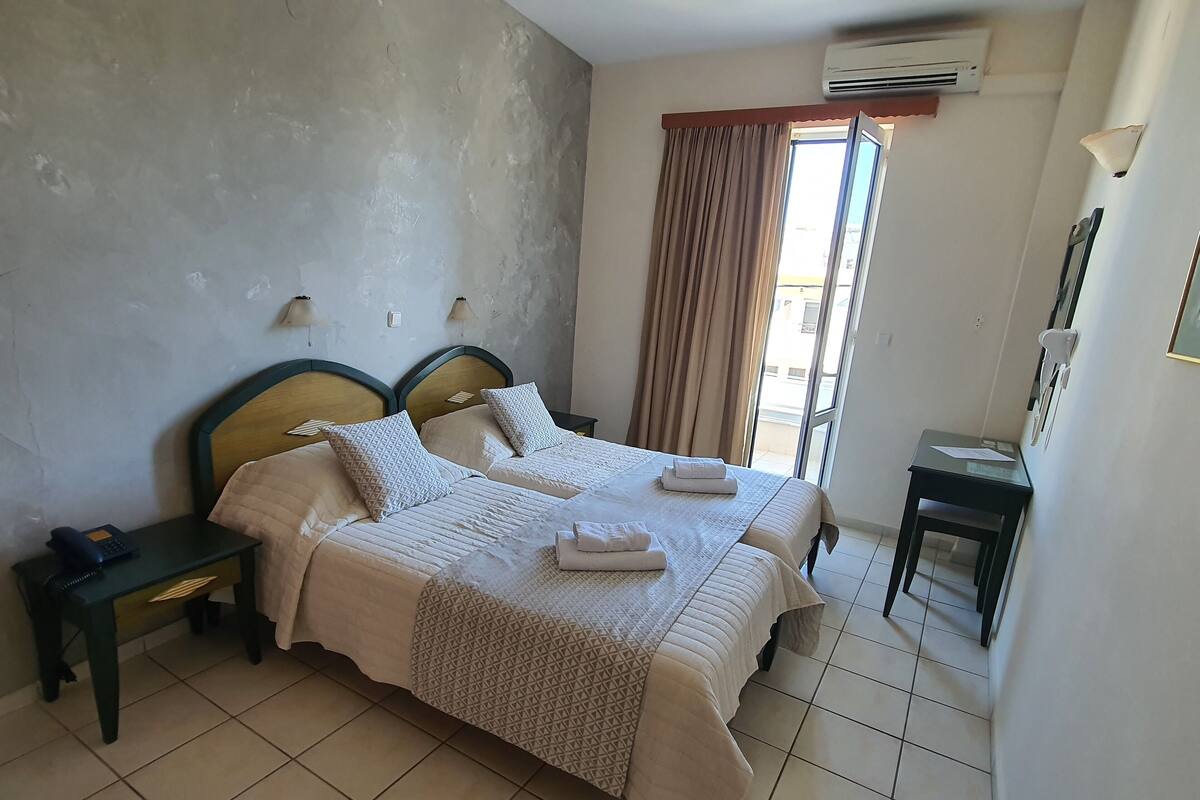 A hotel room with two beds, a bedside table with a phone, a small table with papers, and a view of the outside through a window with curtains.
