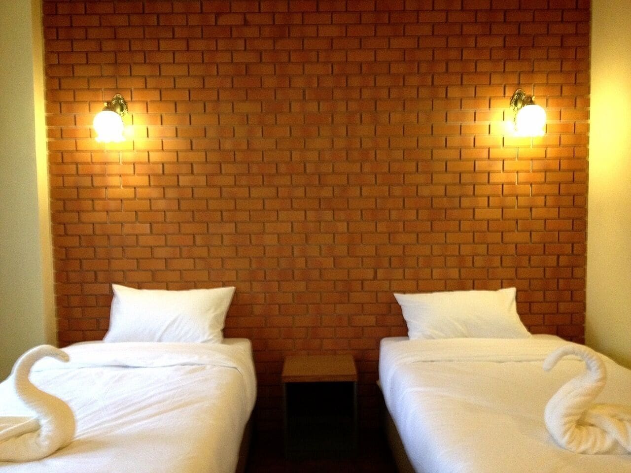 A hotel room with two beds, each with a white bedspread and pillows, and a brick-patterned wall.