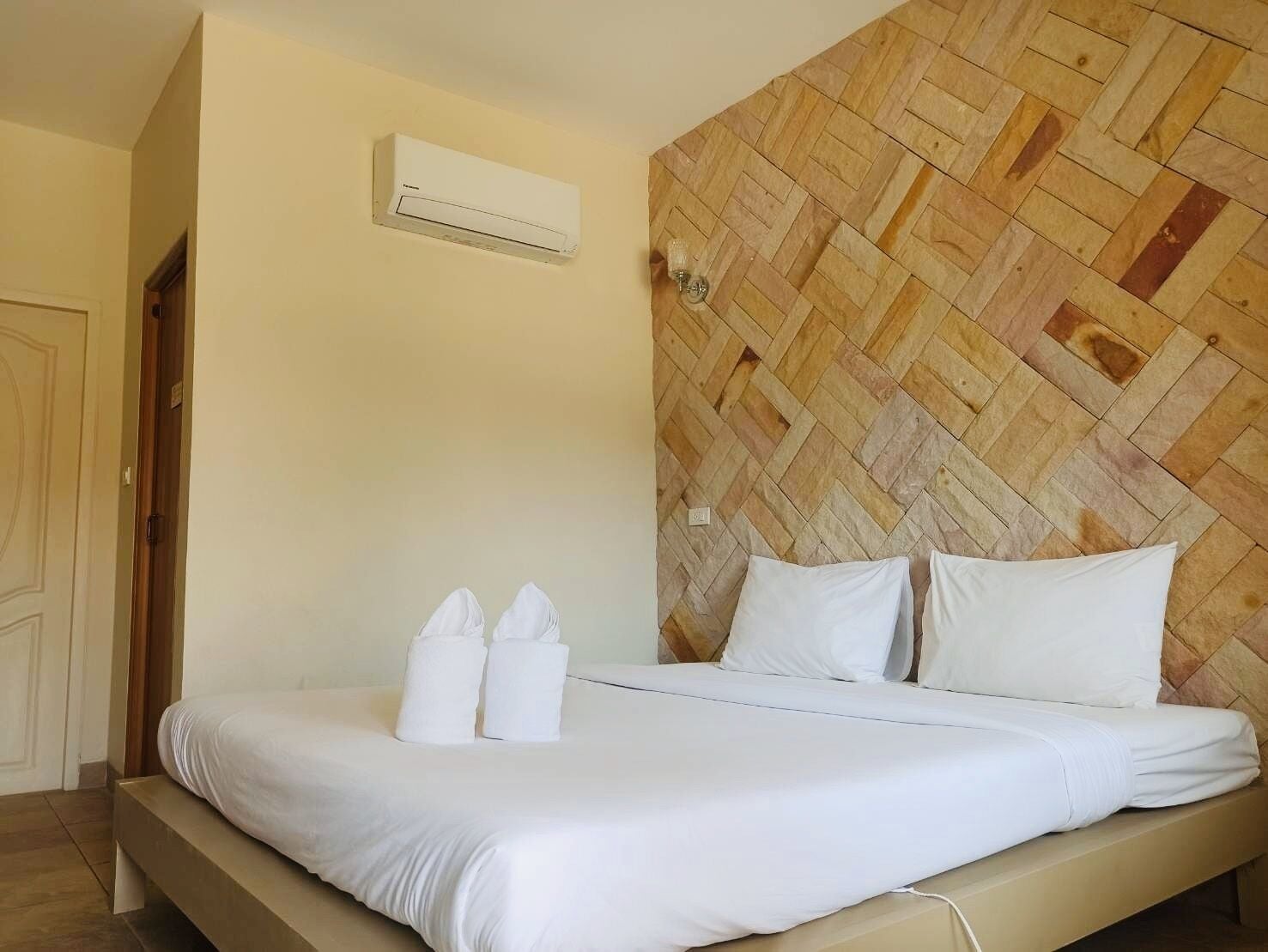 Habitación de hotel con cama, ropa de cama blanca, cabecera de madera, unidad de aire acondicionado y una puerta.