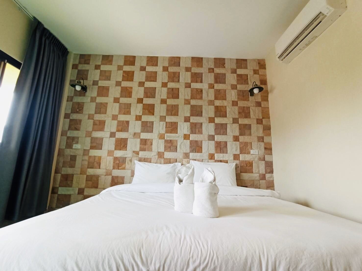 Una habitación de hotel con un estampado de cuadros en la pared, una cama con ropa blanca, dos lámparas fijadas a la pared y un equipo de aire acondicionado.