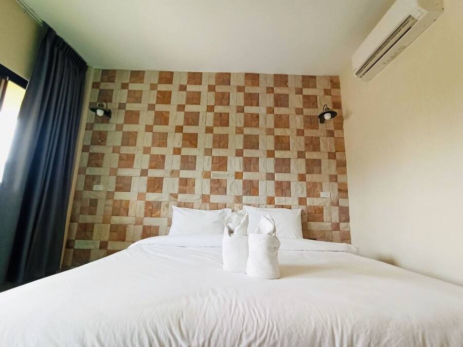 Una habitación de hotel con un estampado de cuadros en la pared, una cama con ropa blanca, dos lámparas fijadas a la pared y un equipo de aire acondicionado.