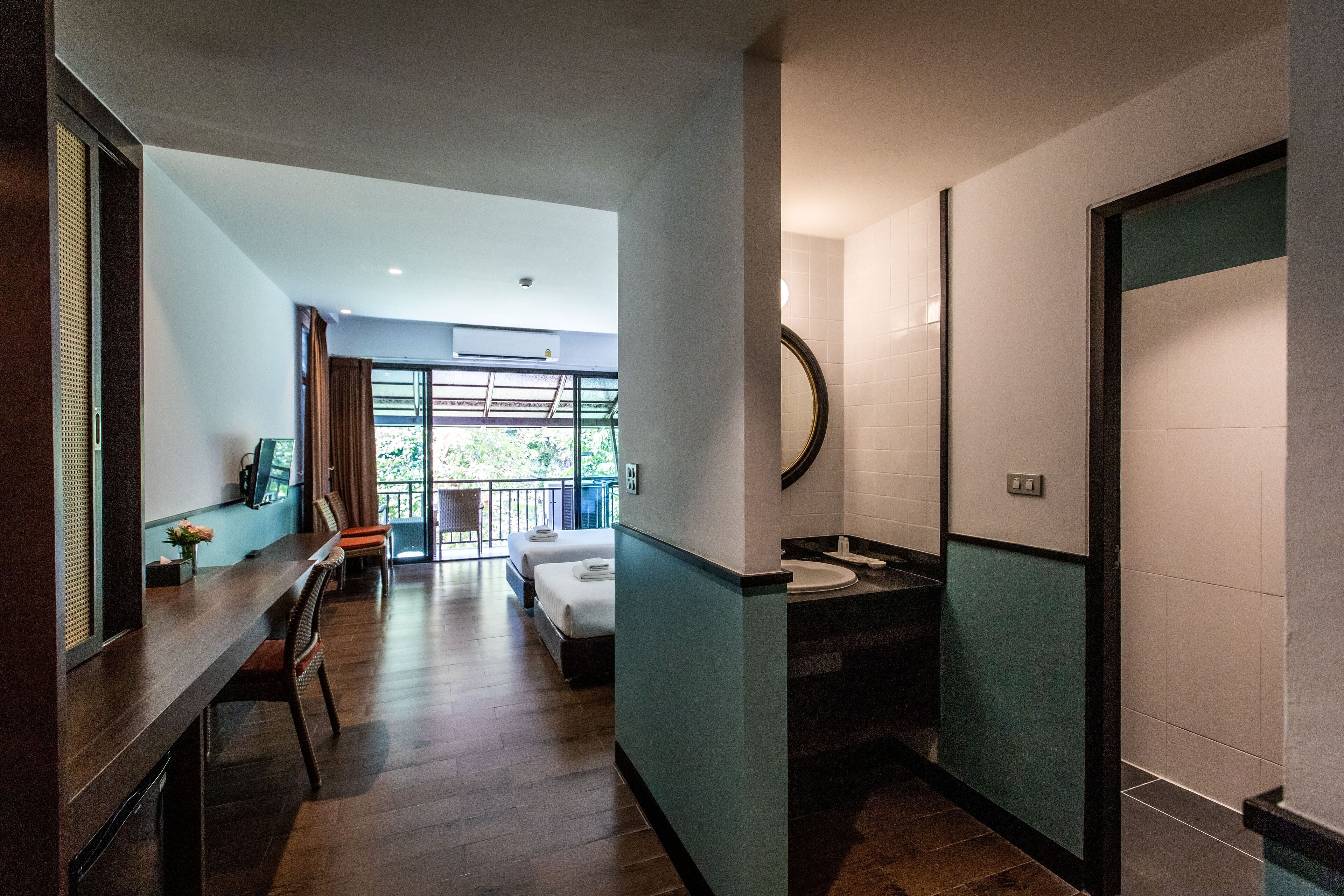A hotel room with a bed, a desk, a chair, a bathroom with a sink and mirror, and a balcony with a view of greenery.