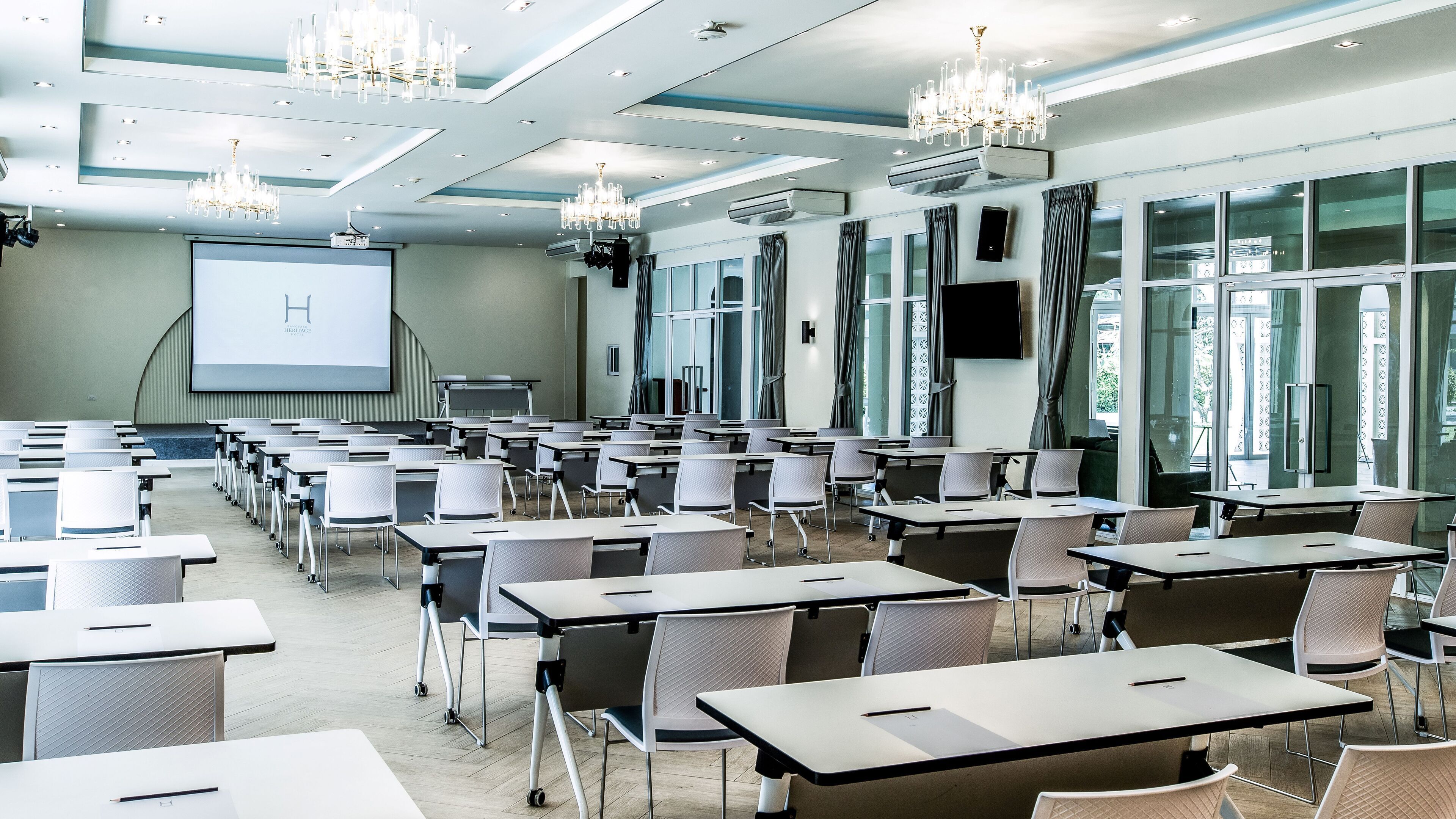 A conference room with rows of tables and chairs, a projector screen, and a large window with curtains.