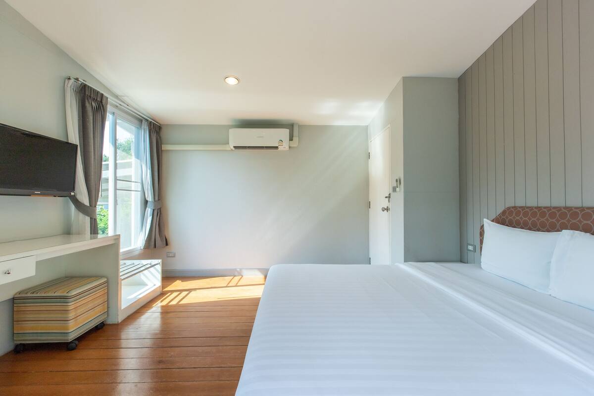 Une chambre d’hôtel équipée d’un lit, d’une télévision, d’une fenêtre avec des rideaux, d’un climatiseur et d’un plancher en bois.