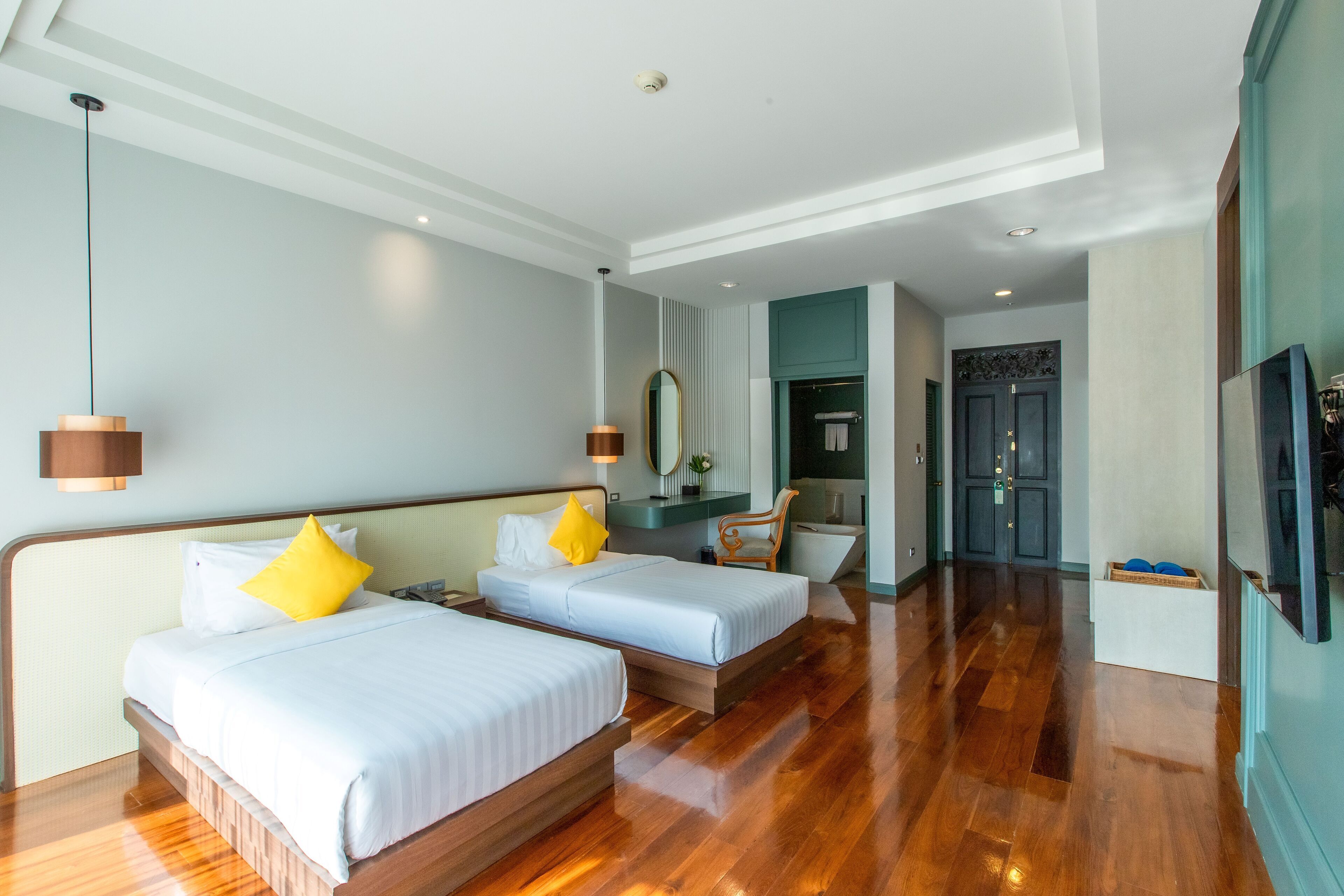A hotel room with two beds, a wooden floor, a mirror, a TV, and a door leading to a bathroom.