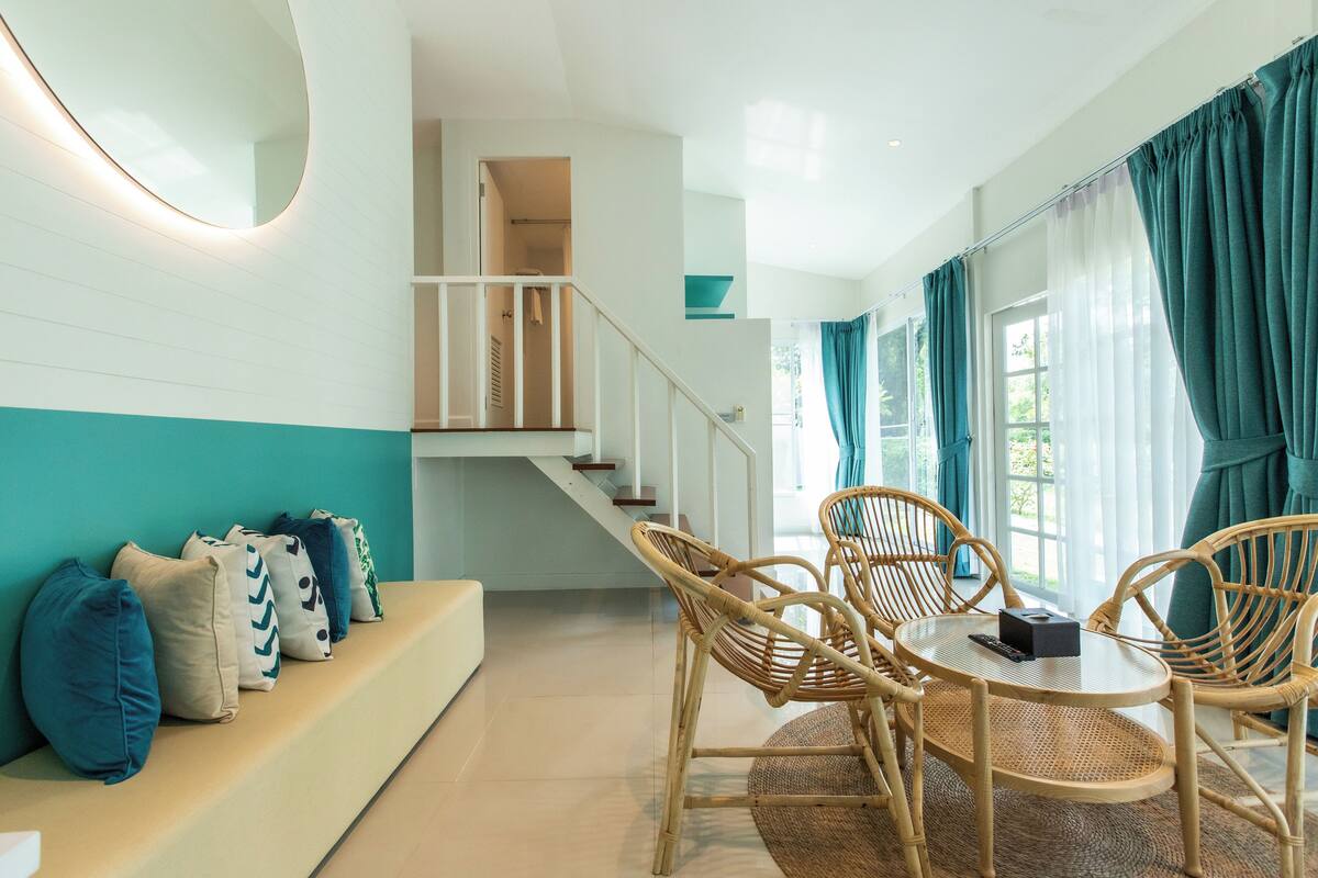 Une chambre d’hôtel moderne avec un canapé, une table à manger avec des chaises, un escalier et de grandes fenêtres équipées de rideaux.