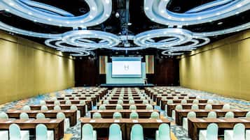 Un salón de conferencias con filas de mesas y sillas, una pantalla grande al frente y un diseño moderno del techo.