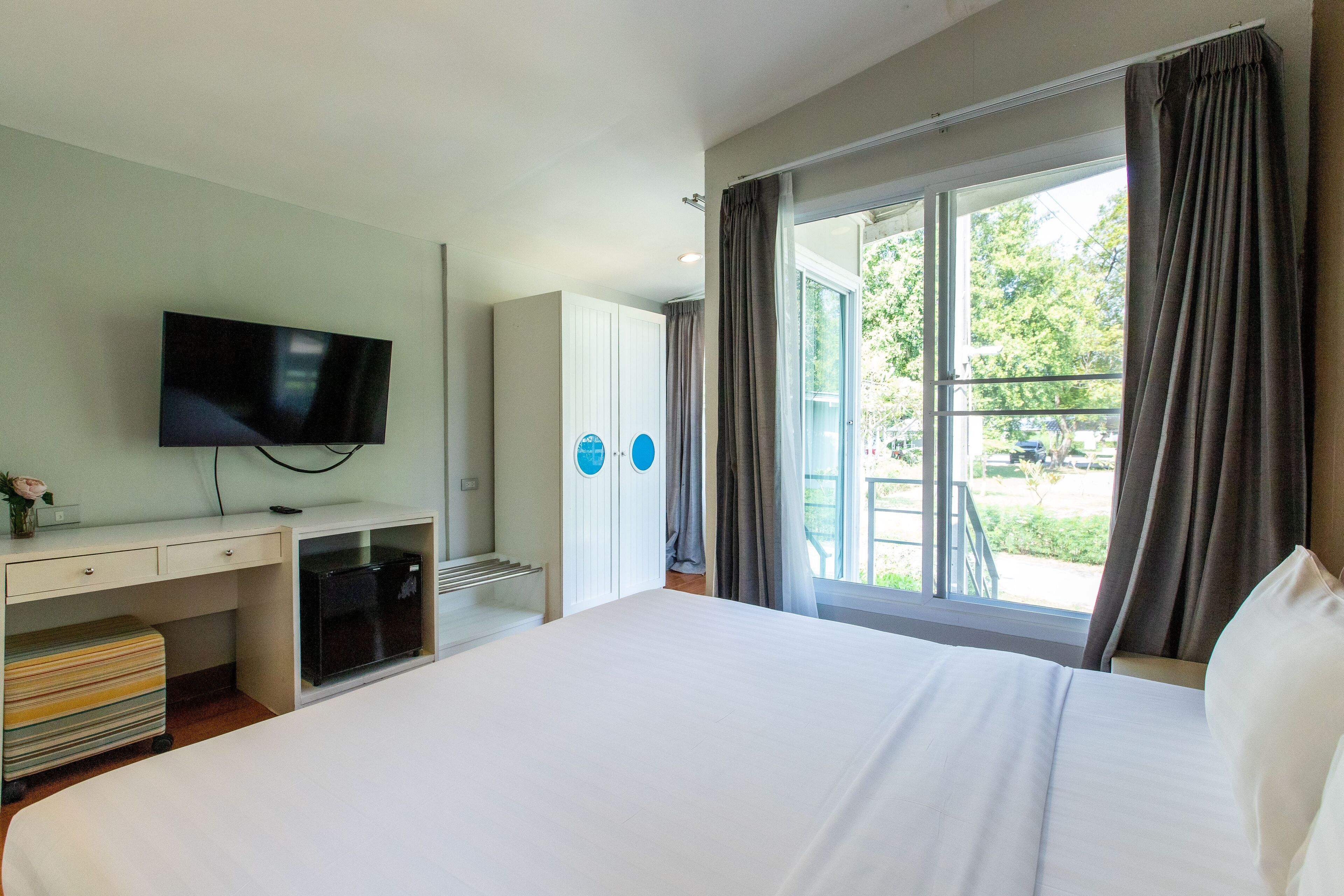 A hotel room with a bed, a desk with a flat-screen TV, a wardrobe, and a large window with curtains.