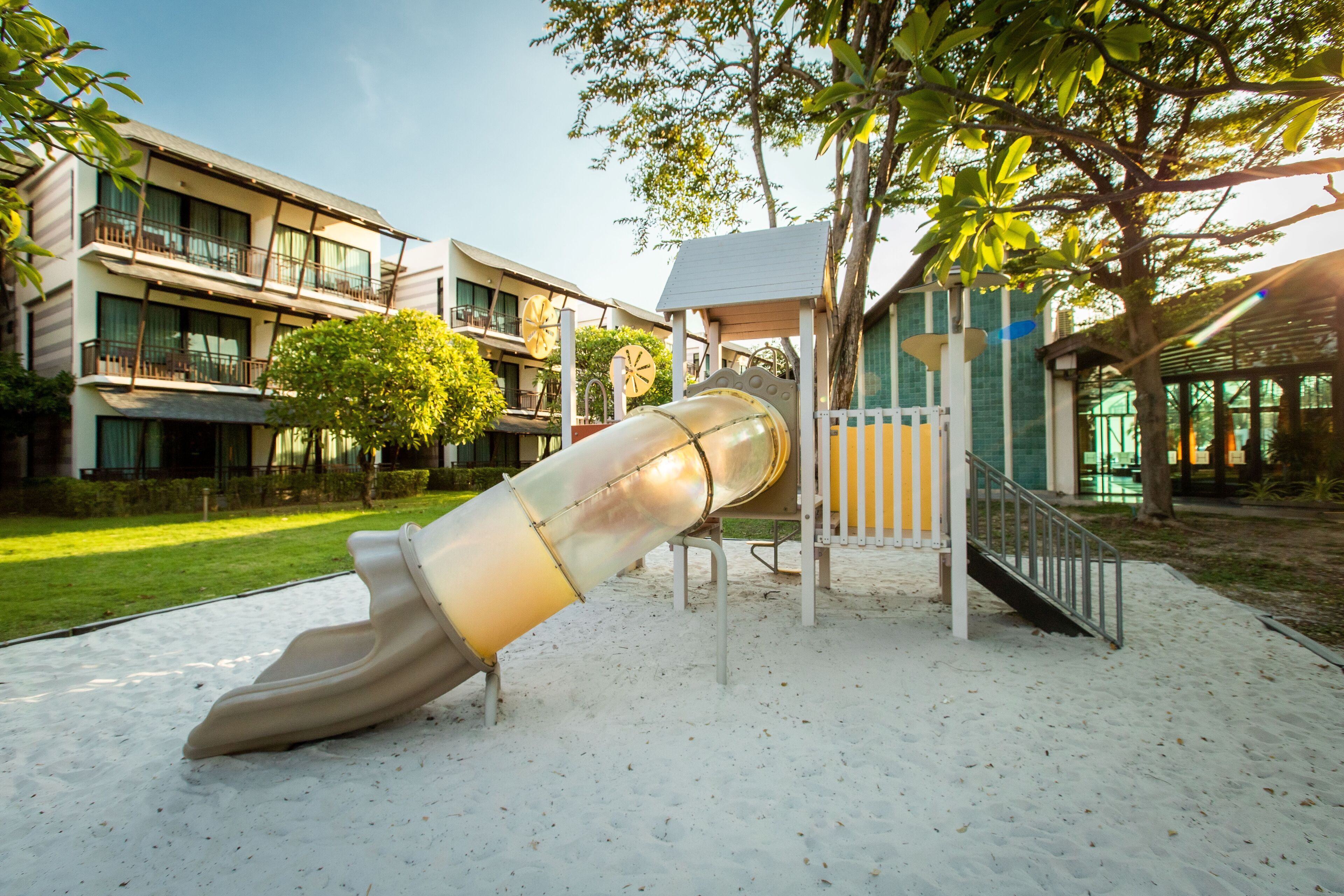 A playground with a slide and climbing structures, surrounded by a building with multiple balconies and a garden area.