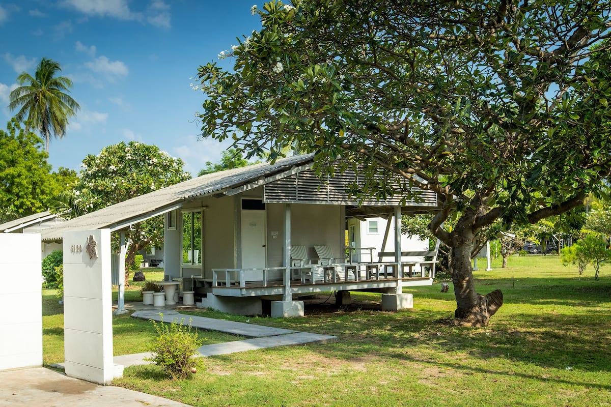 Un petit bâtiment d’un seul étage, doté d’un porche et d’un espace extérieur aménagé avec des sièges, entouré d’une végétation luxuriante et d’arbres.