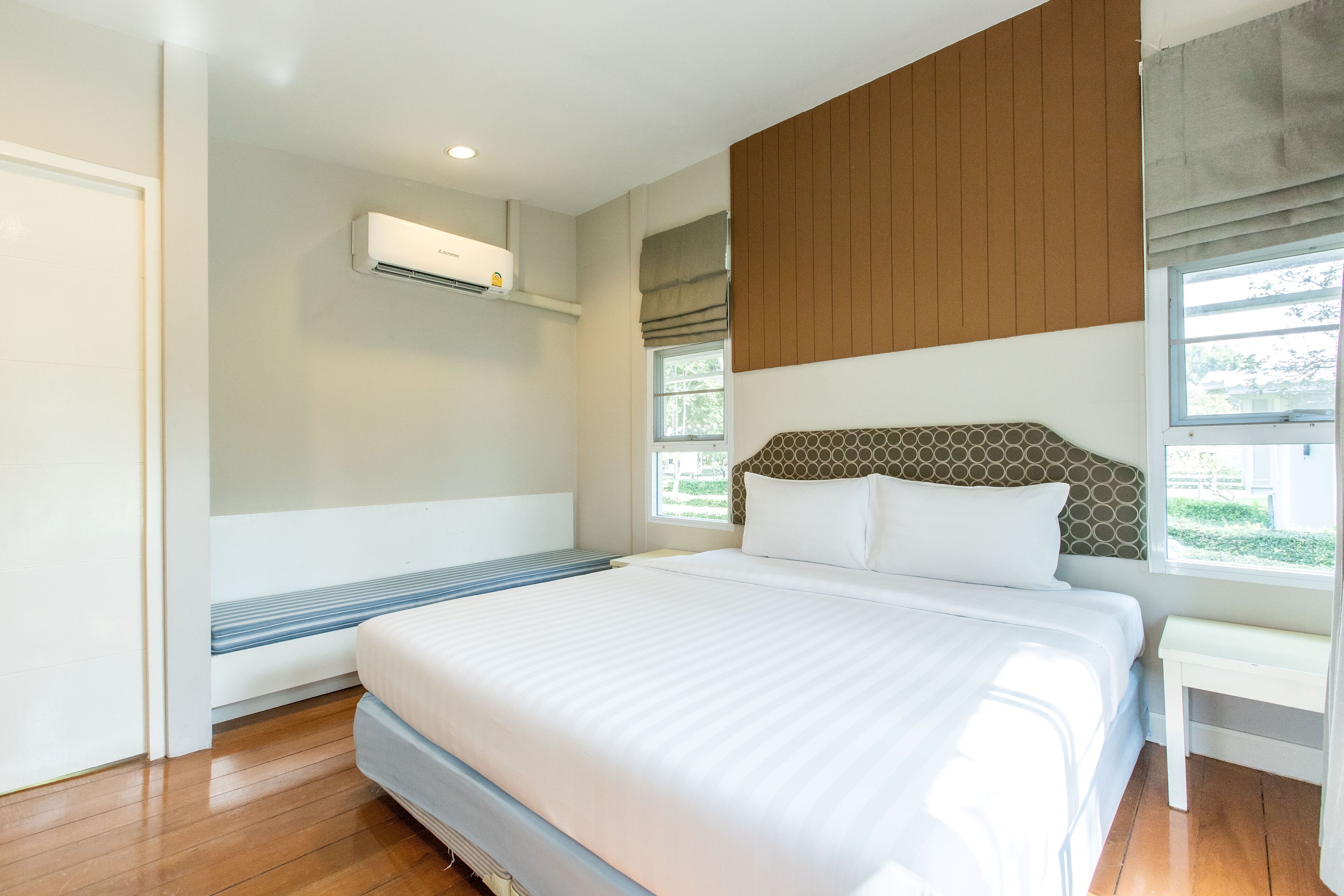 A modern bedroom with a large bed, a built-in bench, an air conditioning unit, and a window with a view of greenery.