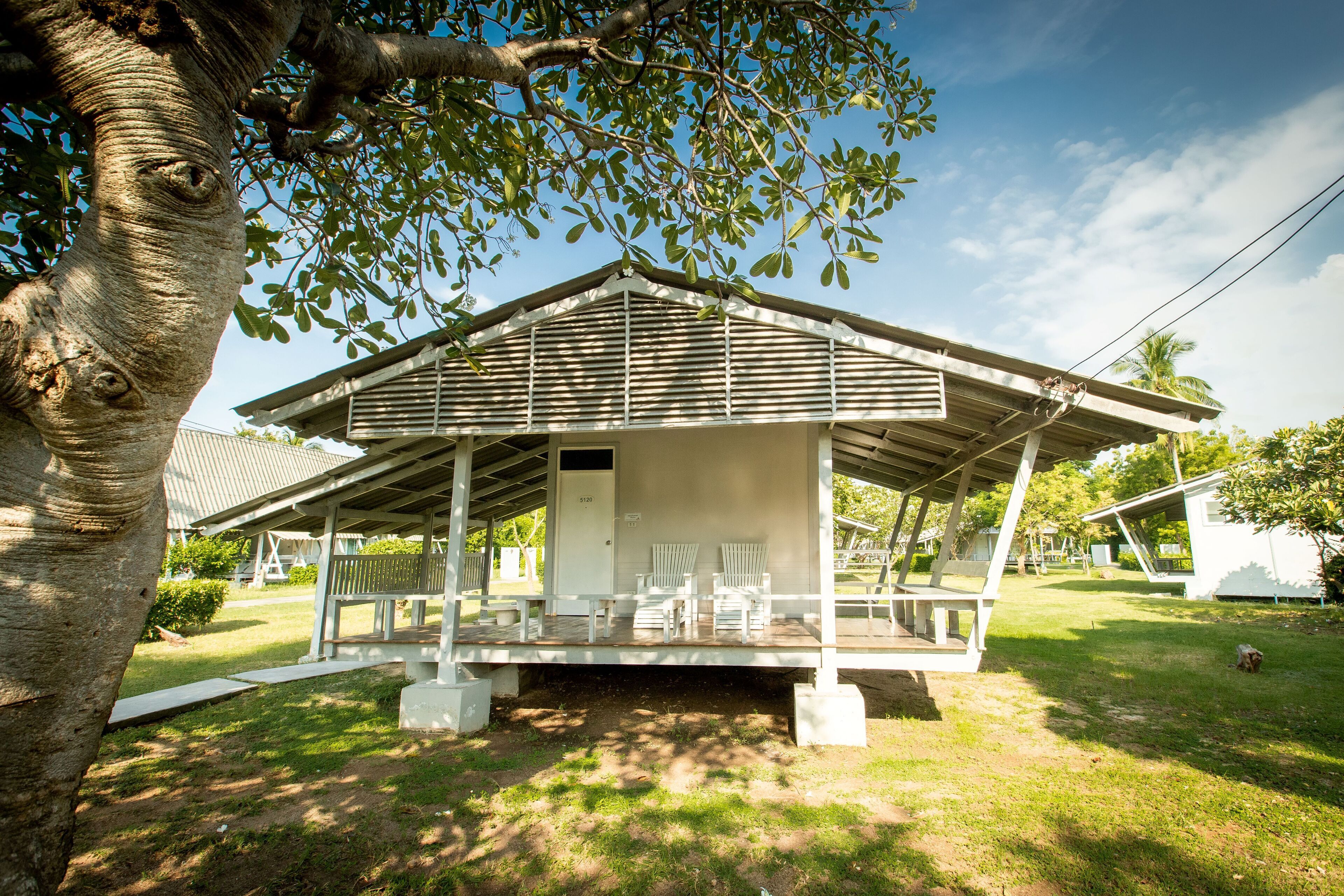 Un espacio de estar al aire libre cubierto, con sillas blancas y un pequeño edificio de techo inclinado.