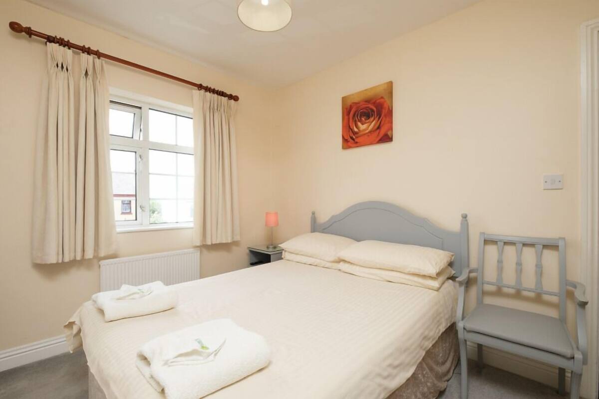 A bedroom with a bed, a chair, a nightstand with a lamp, a window with curtains, and a painting of a rose.
