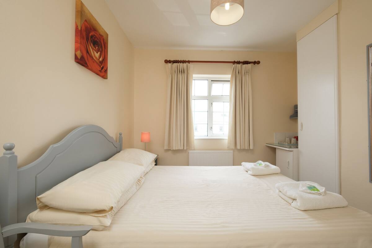 A neatly made bed with white linens, a bedside lamp, and a window with curtains.