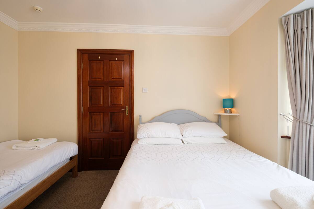 A hotel room with a wooden door, a bed with white bedding, a nightstand with a lamp, and a window with curtains.