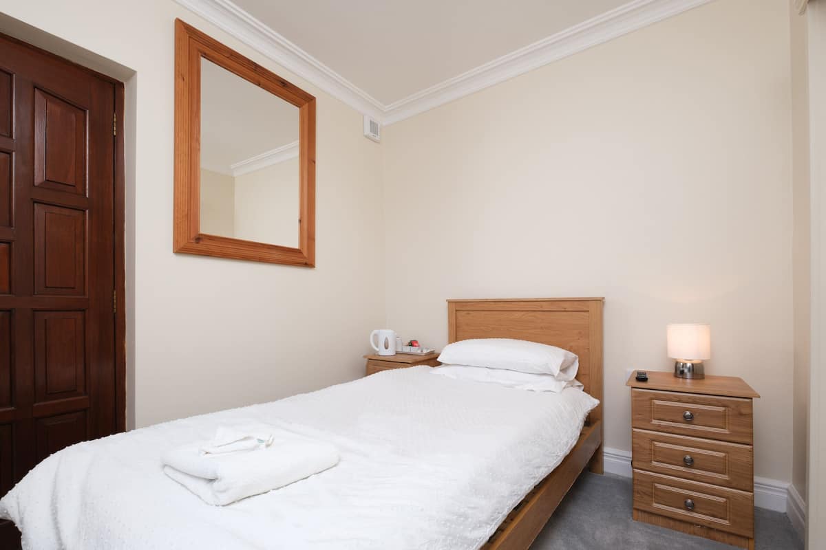 A neatly made bed with a white comforter and a folded towel, a wooden nightstand with a lamp, a wooden wardrobe, and a large mirror on the wall.