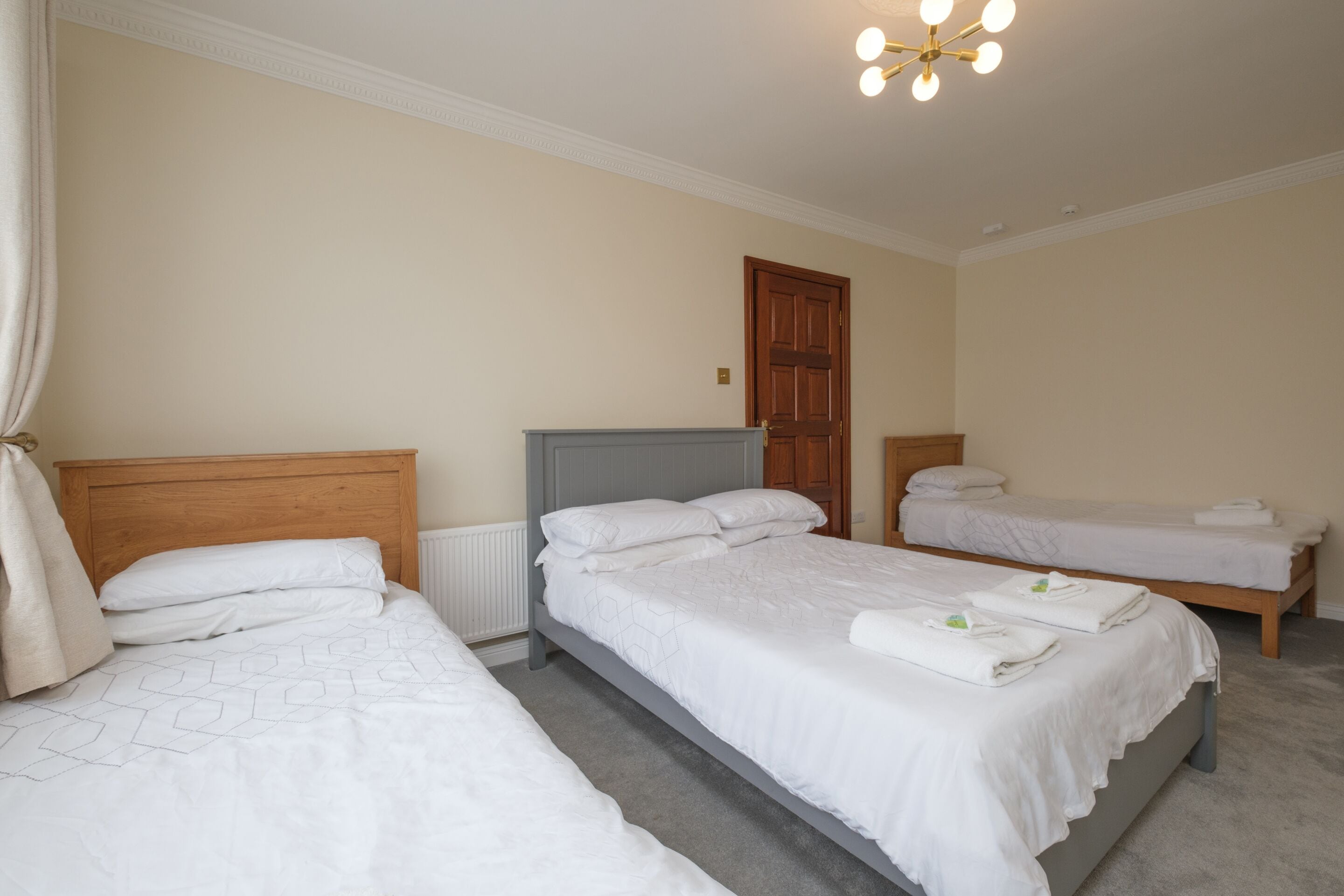 Ein Hotelzimmer mit drei Einzelbetten, einem Holzkopfteil, einem Heizkörper und einer Deckenleuchte.