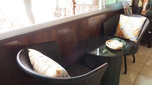 A wicker chair with a floral cushion, a wicker table with a basket, and a view of the outdoors through a window. - Phutawan Kan Resort (Kanchanaburi)