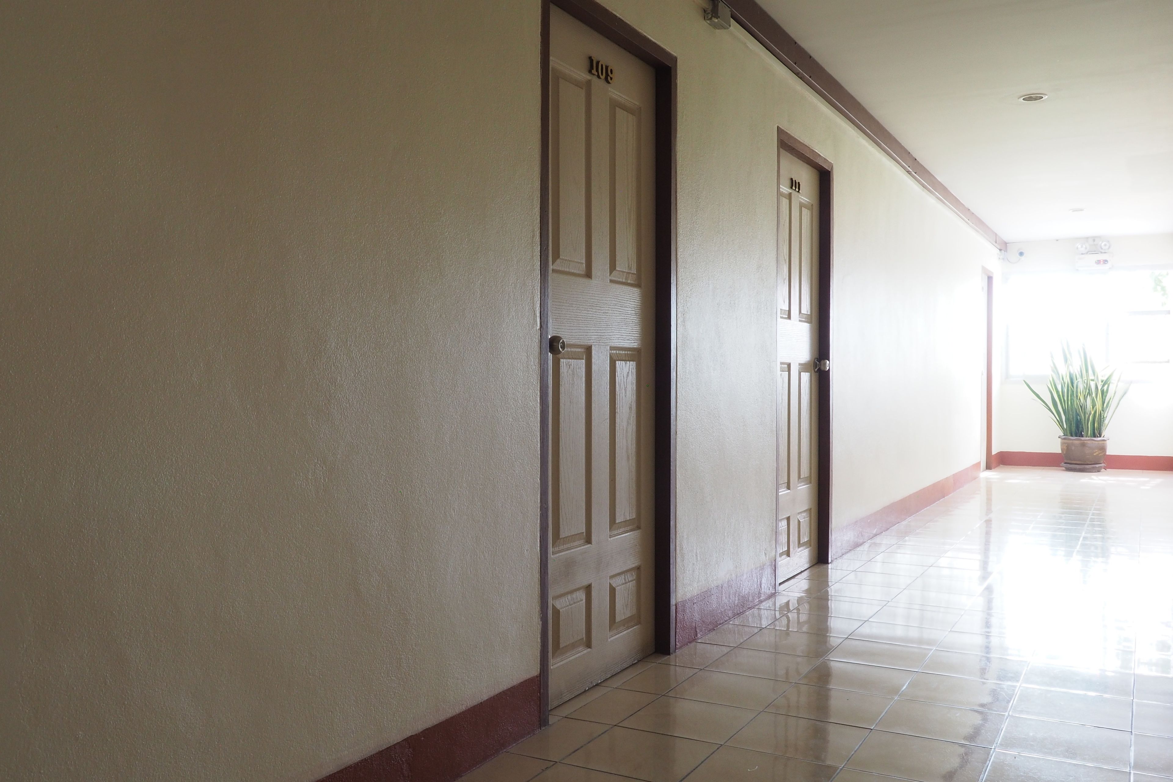 Um corredor com duas portas, uma delas com a placa '101' e a outra '102', dando acesso a quartos. O corredor possui um piso de azulejos e um vaso de plantas no canto.