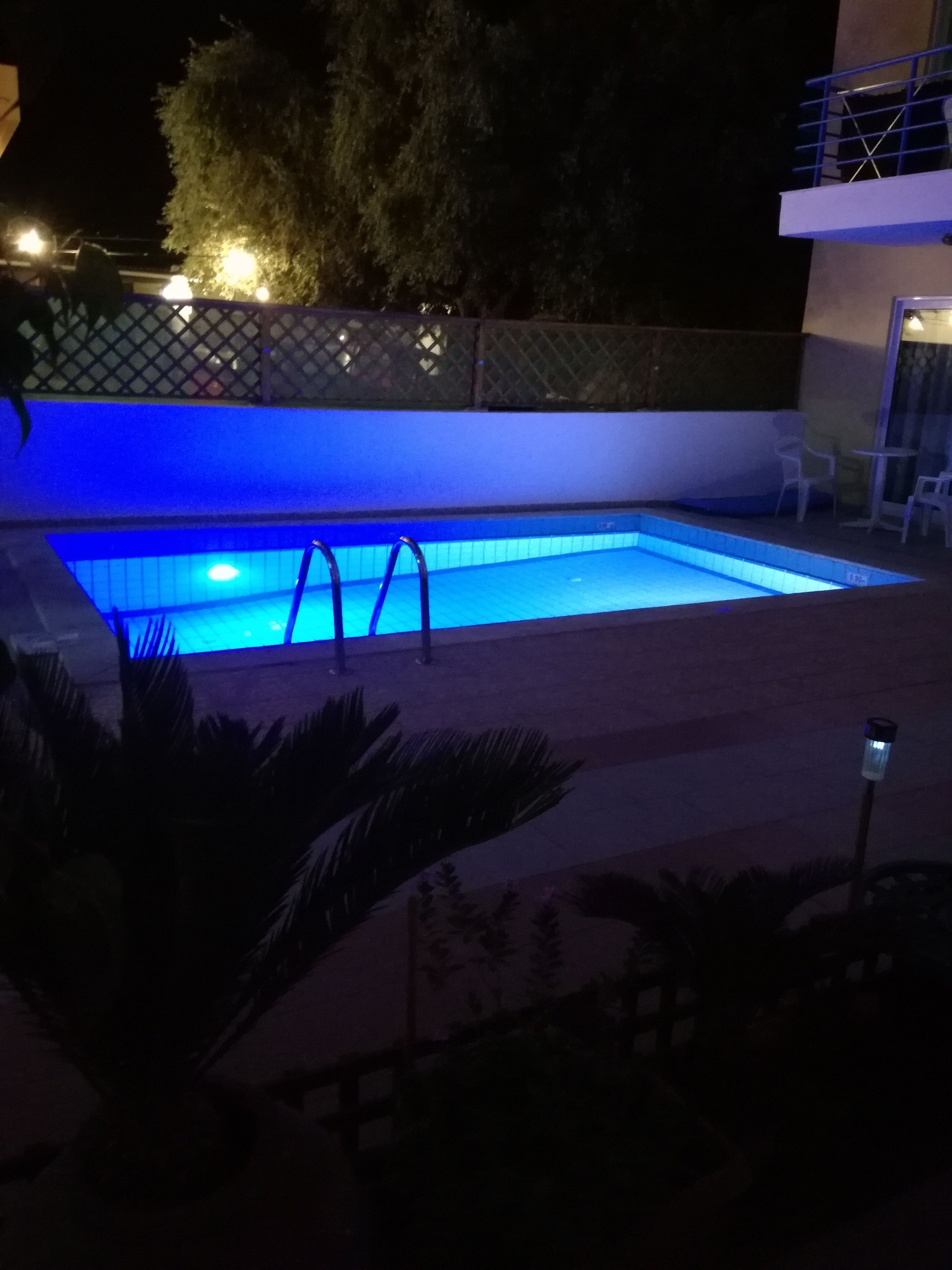 Piscina retangular com borda iluminada em azul, cercada por um muro baixo e uma cerca.