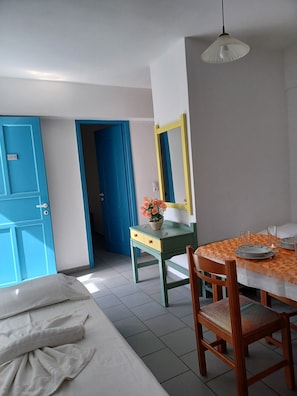 Una habitación de hotel con una cama, un escritorio con un florero, una mesa de comedor con platos y vasos, y un espejo en la pared.