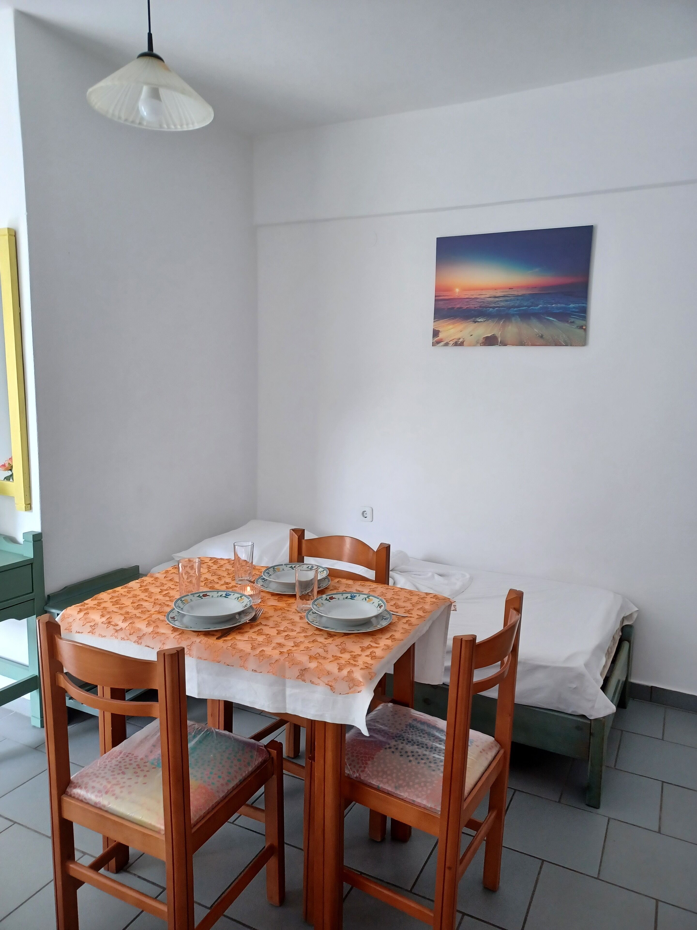 A dining area with a table set for four, chairs, a painting of a sunset, and a white ceiling light.