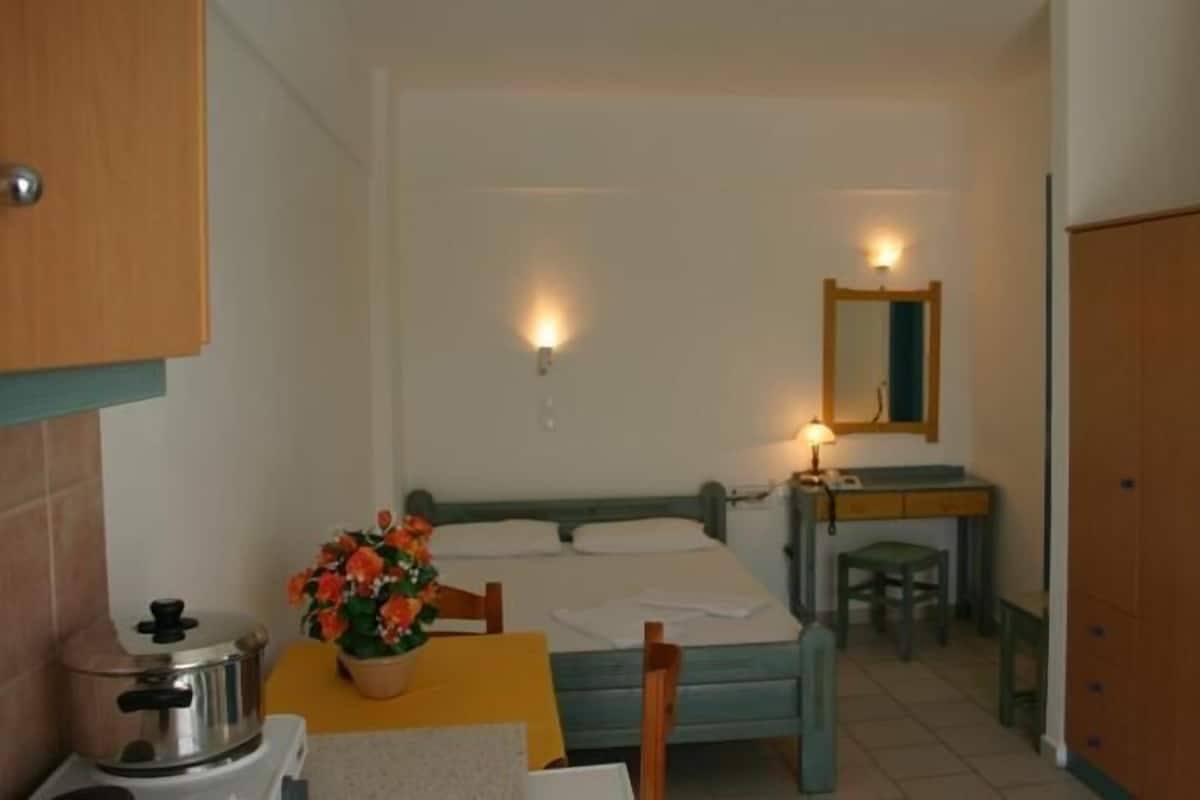 A small, compact room with a bed, a desk, a chair, a table with a flower vase, and a cabinet.