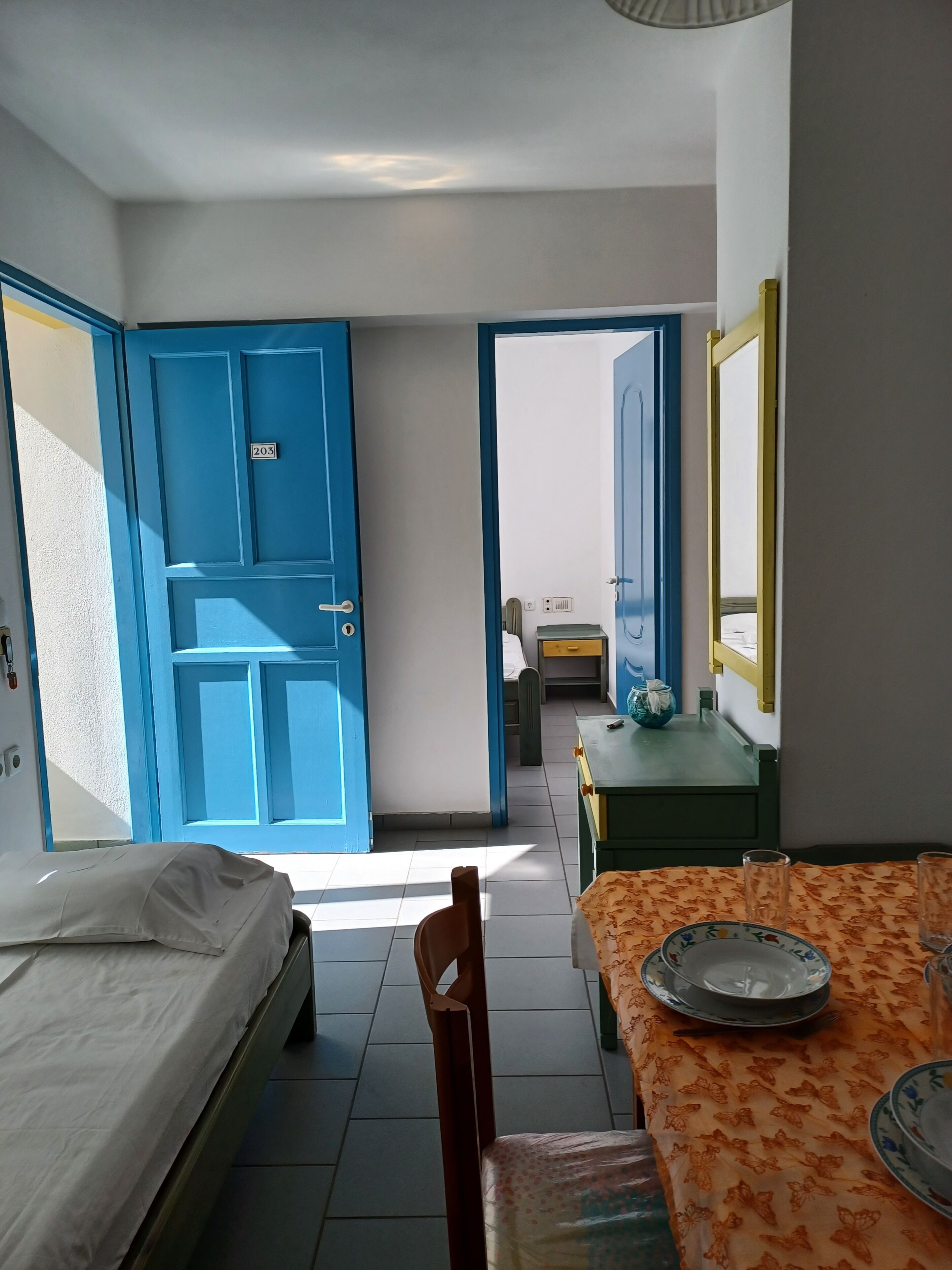 A room with a bed, a table with a floral tablecloth, and a blue door with a number plate.