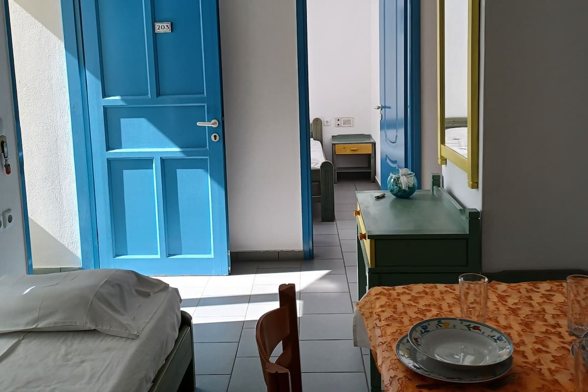 A room with a bed, a table with a floral tablecloth, and a chair. There is a blue door with a number plate, a window, and a mirror on the wall.