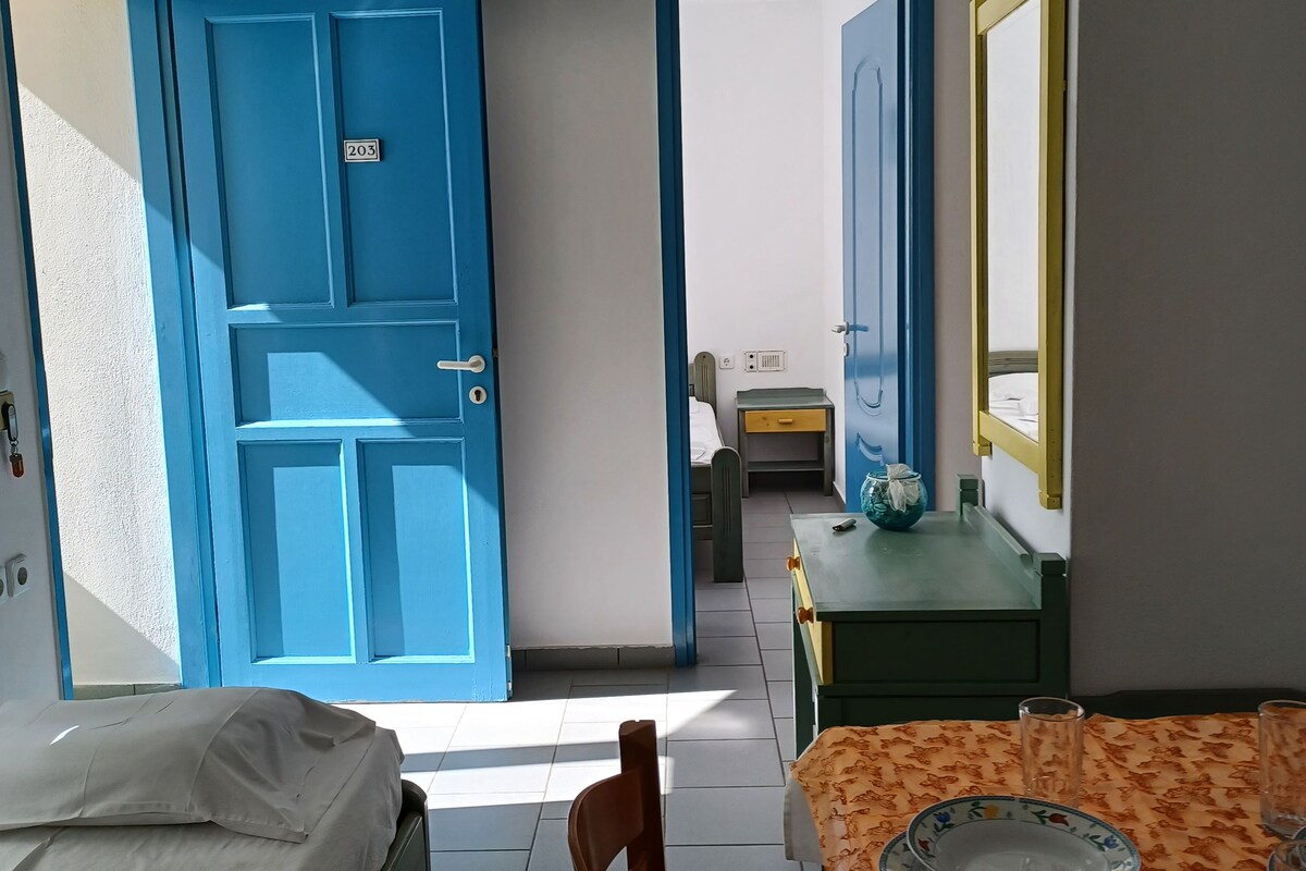 A room with a bed, a table with a floral tablecloth, and a chair. There is a blue door with a number plate, a window, and a mirror on the wall.