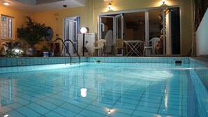 An indoor swimming pool with tiled flooring, a pool ladder, and a seating area with chairs and tables. - Olympic Star Apartments (Hersonissos)