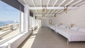 Una habitación amplia y luminosa, con grandes ventanales, una cama larga de color blanco y vista al mar.