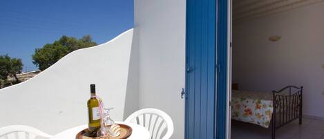 A small balcony with a white table, a bottle of wine, and two glasses. The balcony door is blue, and there is a bed with a floral bedspread inside the room.