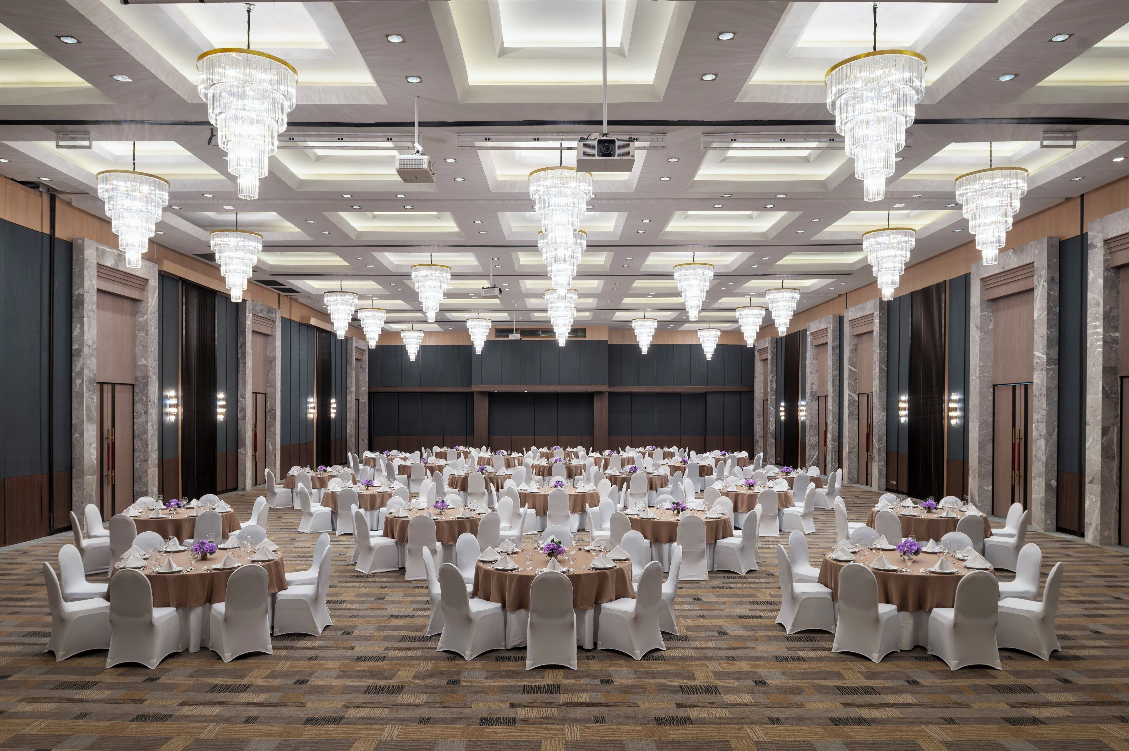 A banquet hall with round tables, white chairs, and chandeliers.