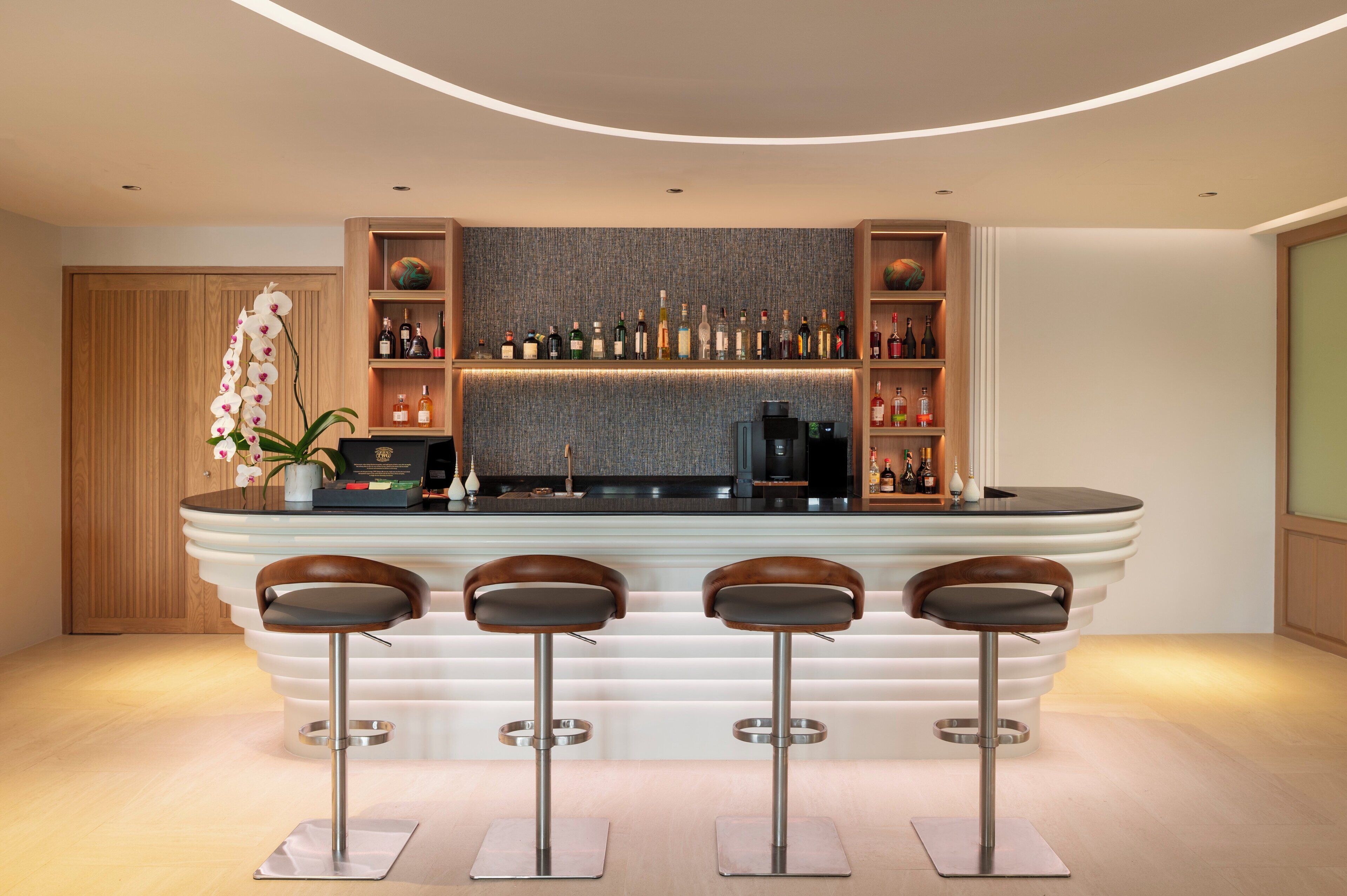 A modern bar area with a curved countertop, three stools, and a variety of bottles and glasses on shelves.