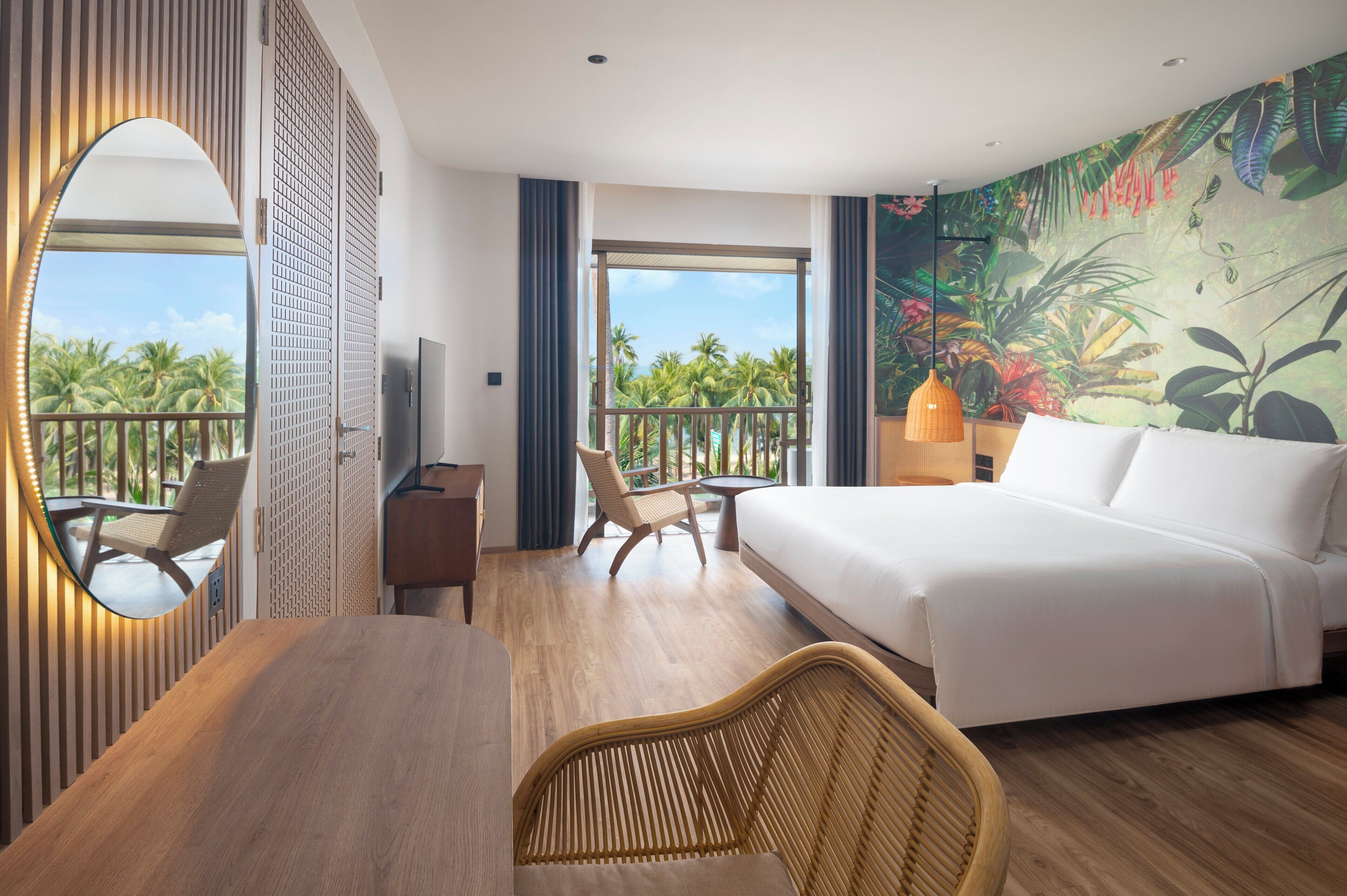 Ein Hotelzimmer mit einem großen Bett, einem Schreibtisch mit Stuhl, einem kleinen Tisch, einem Fernseher und einem Balkon mit Blick auf Palmen.