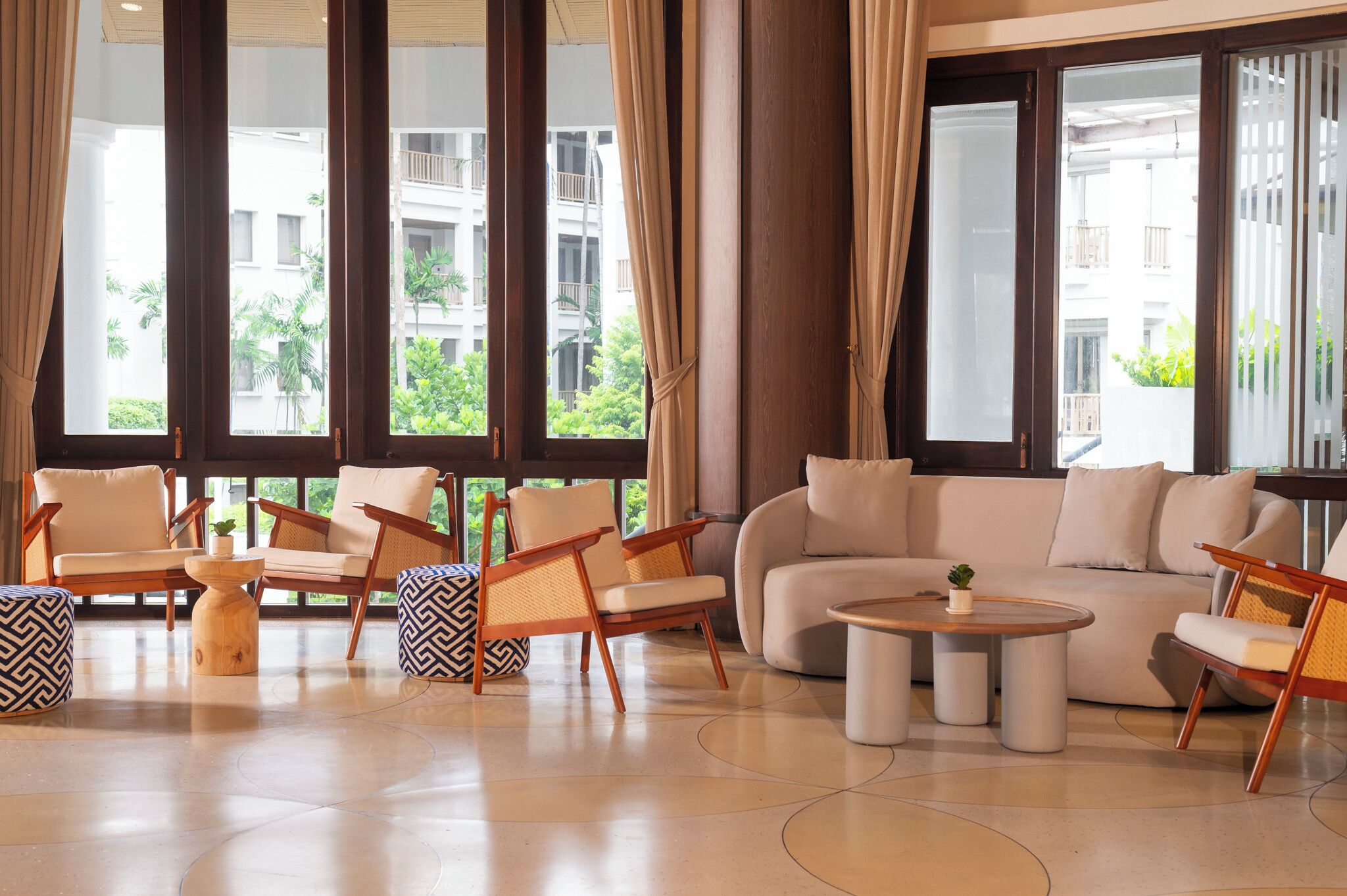 Un vestíbulo amplio con un sofá beige, dos sillones y una mesa de centro. Amplias ventanas ofrecen vistas a zonas verdes y edificios.