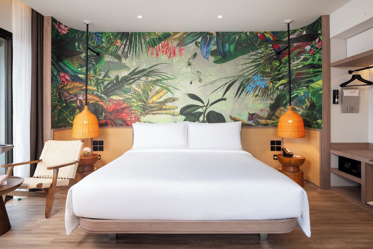 Un dormitorio con una cama grande, dos lámparas de noche, una silla y un mural de follaje tropical.