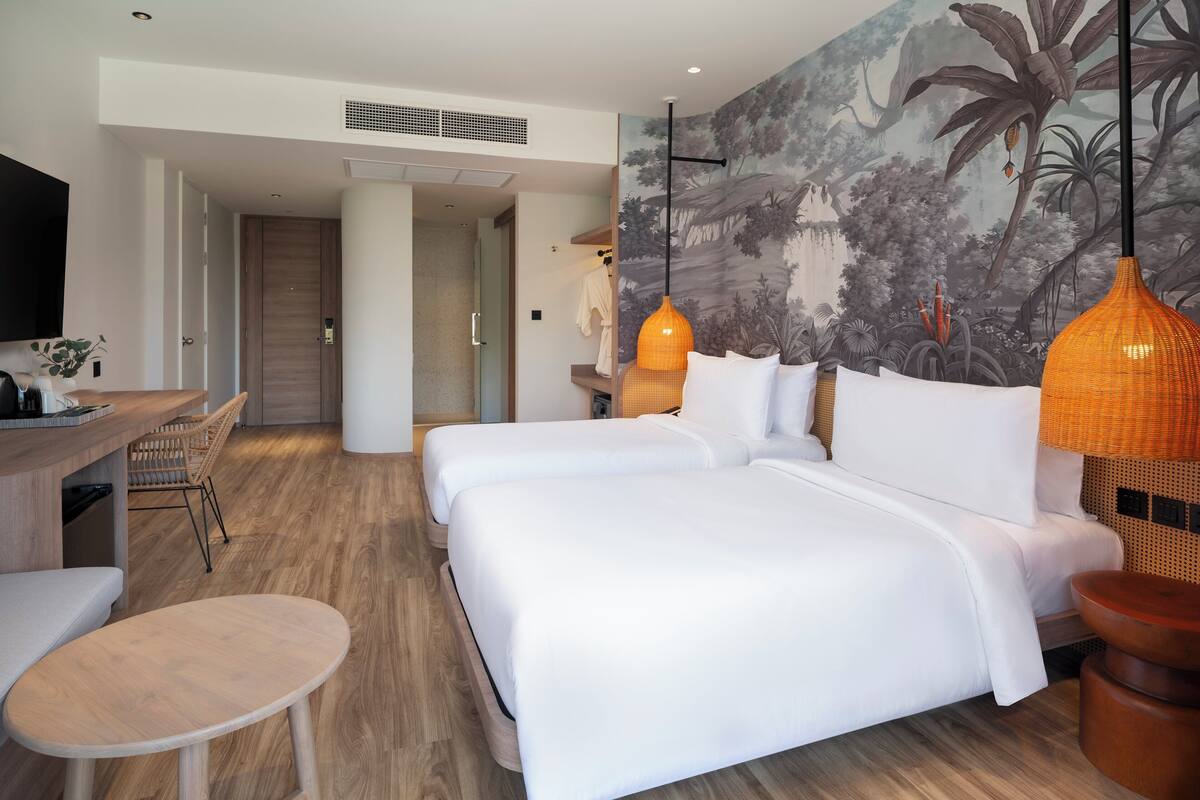 Habitación de hotel con dos camas, un escritorio y una silla. Hay un mural en la pared y una lámpara colgante.