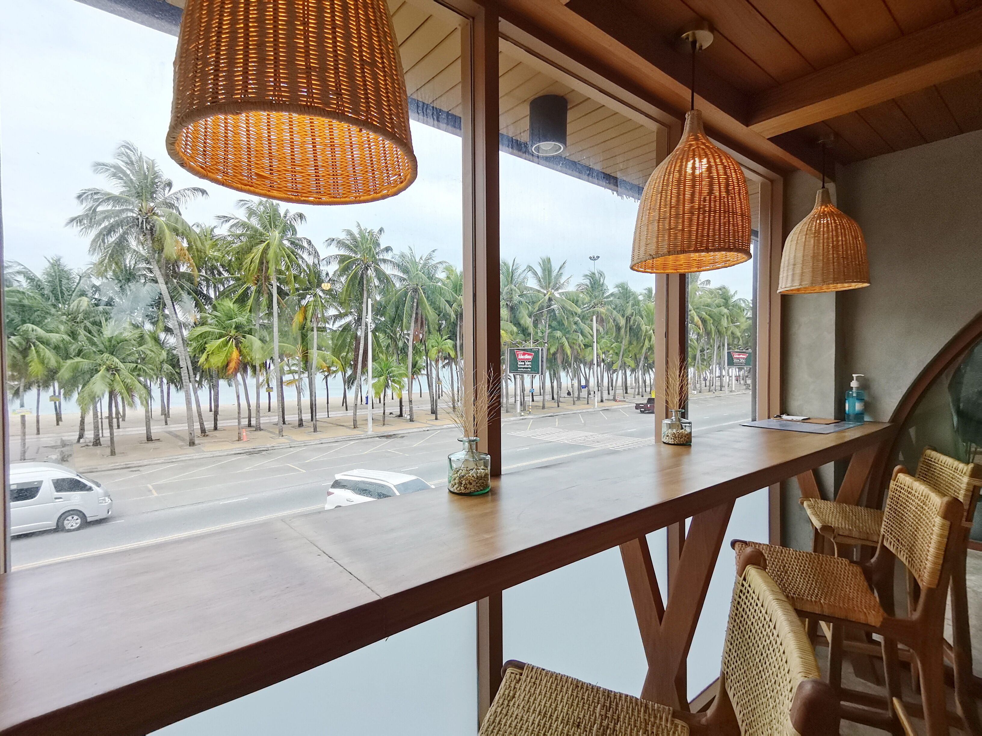 Un restaurant avec des chaises en osier et des lampes suspendues, offrant une vue sur une rue bordée de palmiers et une plage.