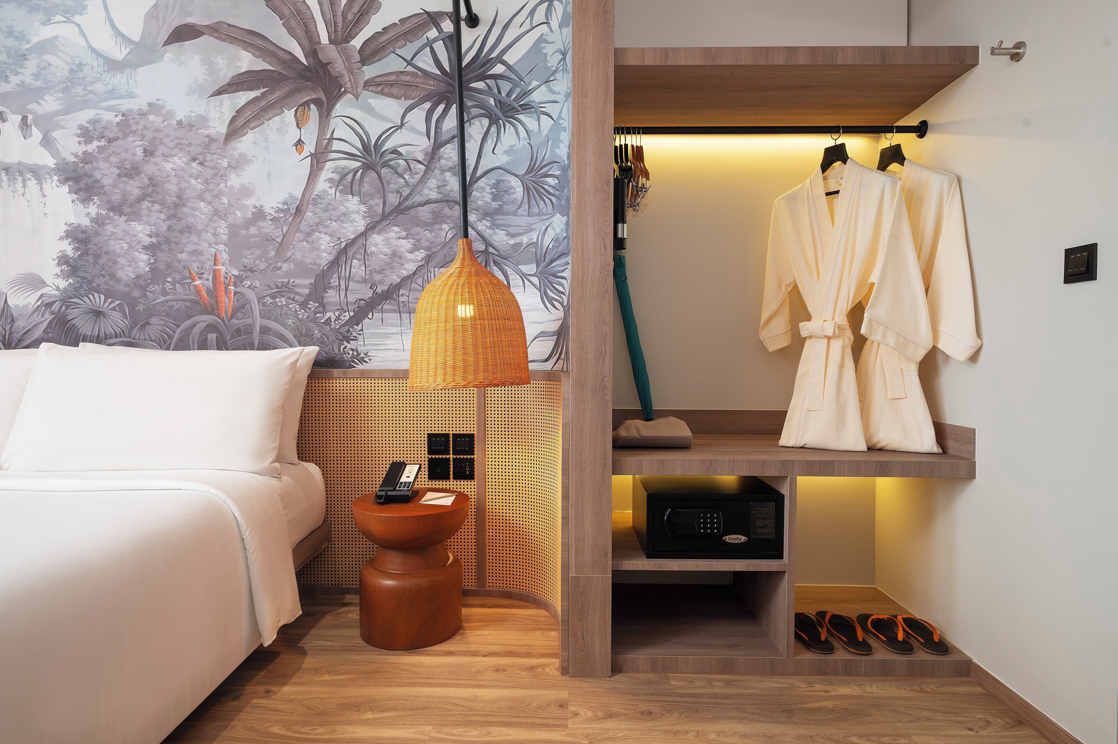 A hotel room with a bed, a wall-mounted telephone, a wooden side table, a robe hanging on a rack, and a shelf with shoes and a radio.