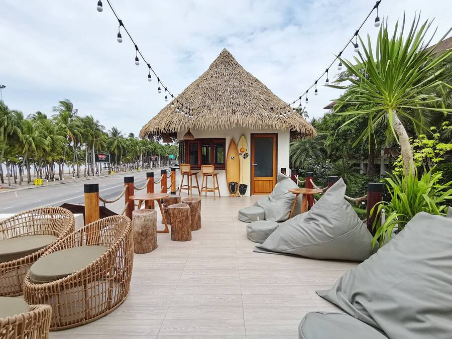A beachside hut with a thatched roof, surrounded by lounge chairs and cushions, and a surfboard leaning against the wall.
