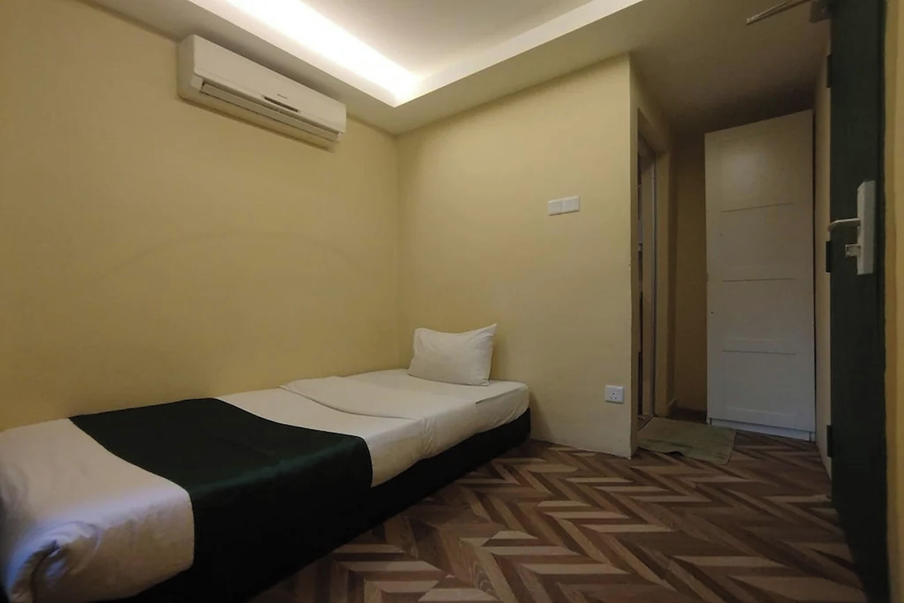 Una habitación pequeña con una cama, un equipo de aire acondicionado, un piso estampado y una puerta que da a otra habitación.