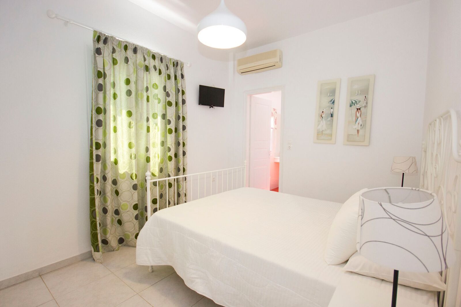 A bedroom with a bed, a white headboard, a curtain, a wall-mounted TV, and a door leading to another room.