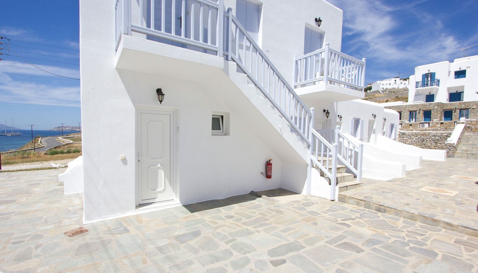A white building with a stone-paved entrance, a fire extinguisher, and a staircase leading to a balcony.