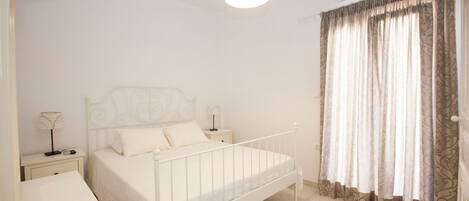 A bedroom with a white bed, a bedside table, a lamp, an air conditioning unit, and a window with curtains.